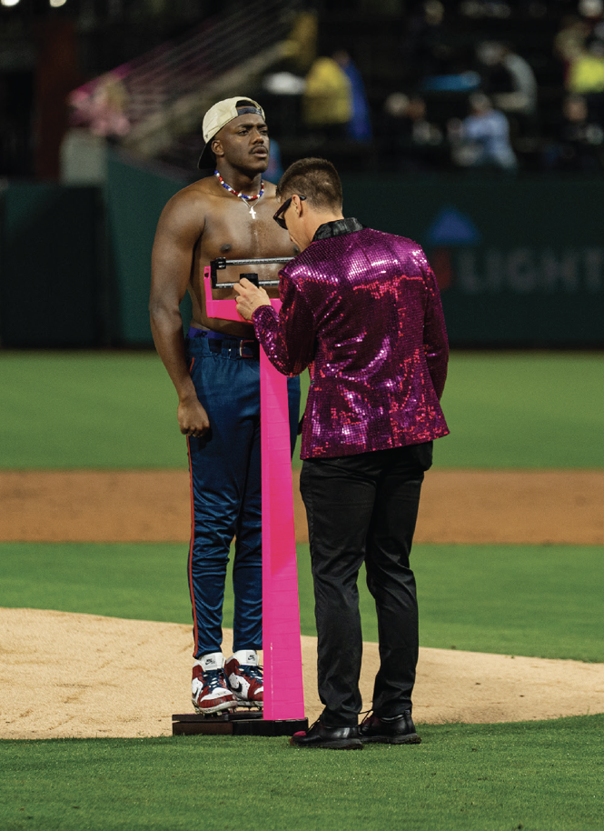 photo of shirtless man in baseball pants, cap, and cleats standing on hot pink doctor's scale being weighed near pitcher's mound by a man in hot-pink-sequined coat and black pants