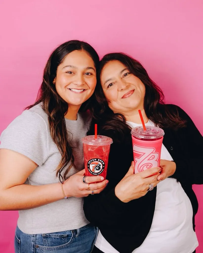 Two women holding 7 Brew drinks against a pink background.