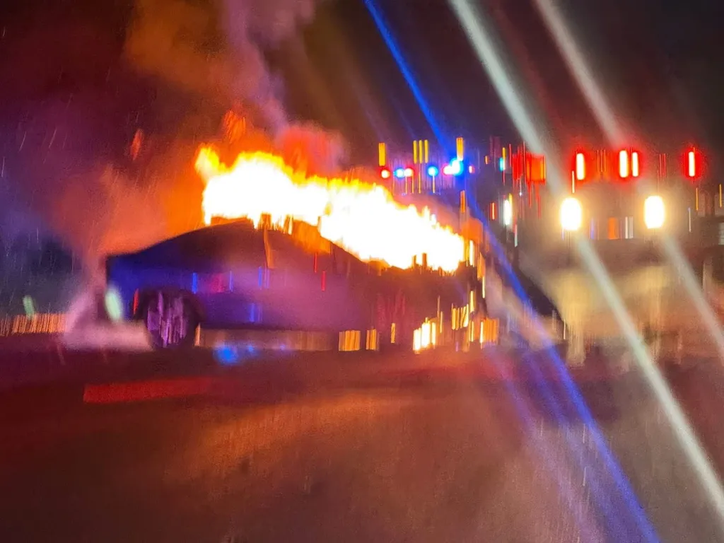 A car engulfed in flames with emergency vehicle lights in the background.