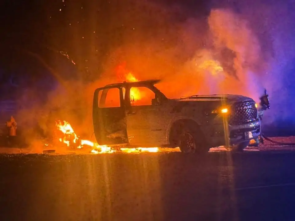 A truck engulfed in flames with a firefighter in the background.