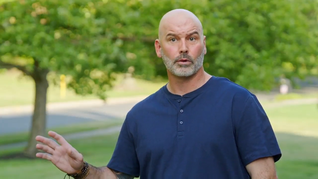 Marissa's ex-husband, Ed, in a blue shirt with his hand raised, outdoors.