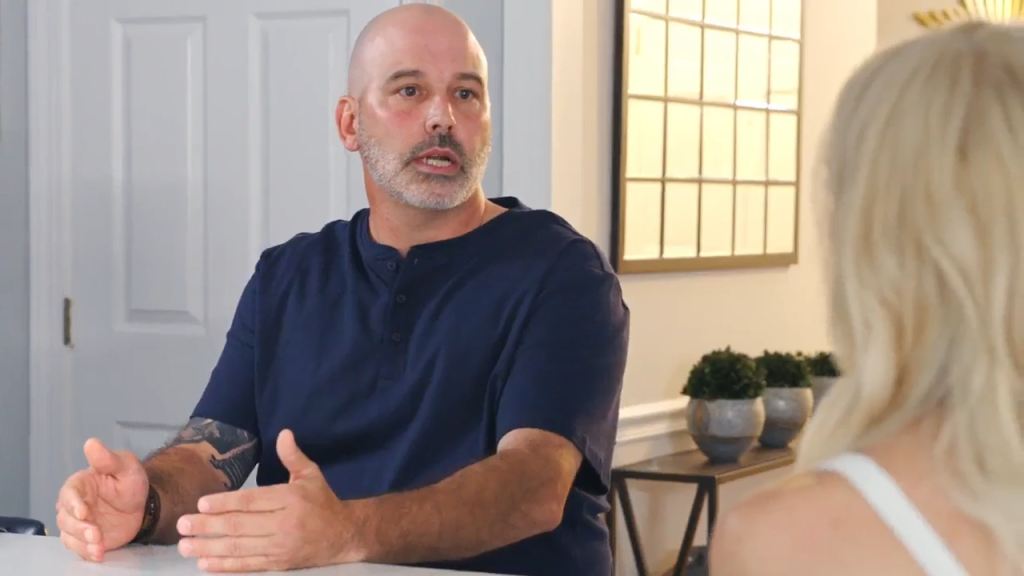 A bald man with a beard and tattoos on his arms, dressed in a navy blue shirt, gestures with his hands while speaking to a blonde woman whose back is to the camera.