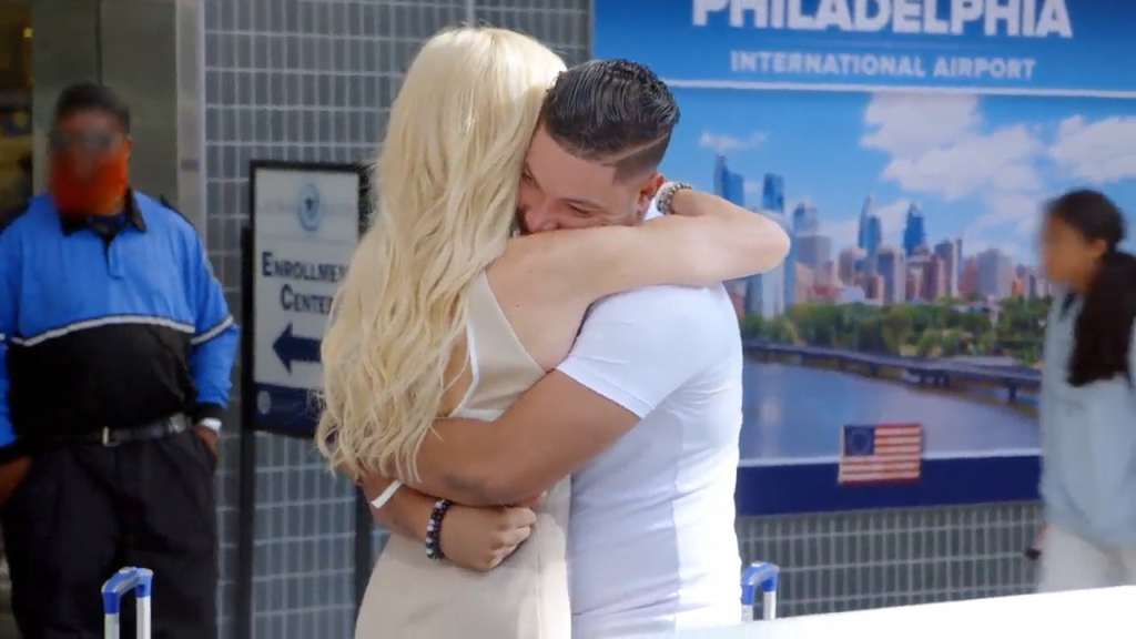 A blonde woman and a man in a white shirt embrace at an airport, with a 