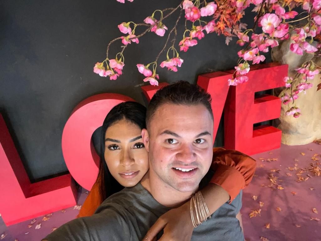 Patrick Mendes and Thais Ramone posing for a selfie in front of red 