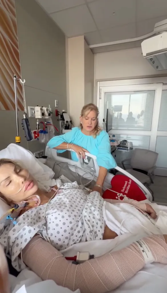 Nikki Exotica in a hospital bed with medical tubes and bandages, with a woman sitting beside her.