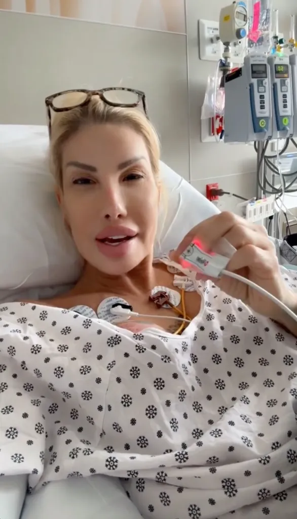 Nikki Exotica in a hospital bed with medical sensors on her chest and holding a pulse oximeter.