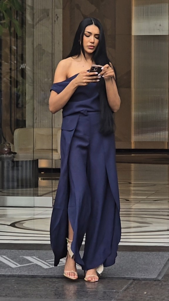 Thais Ramone in a navy blue one-shoulder jumpsuit with wide-leg, draped pants and wrap overlays, looking at her phone.