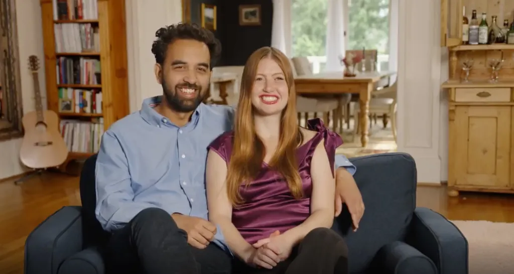 A man and woman sit together on a blue couch, smiling at the camera.