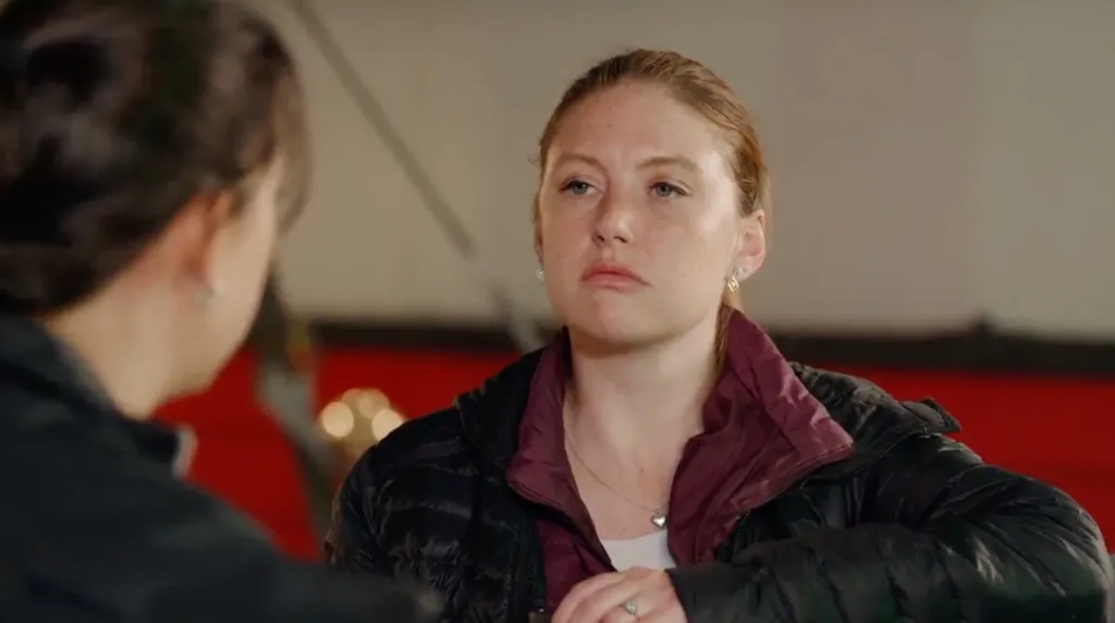 Catie with a serious expression, wearing a black puffer jacket and a heart necklace.