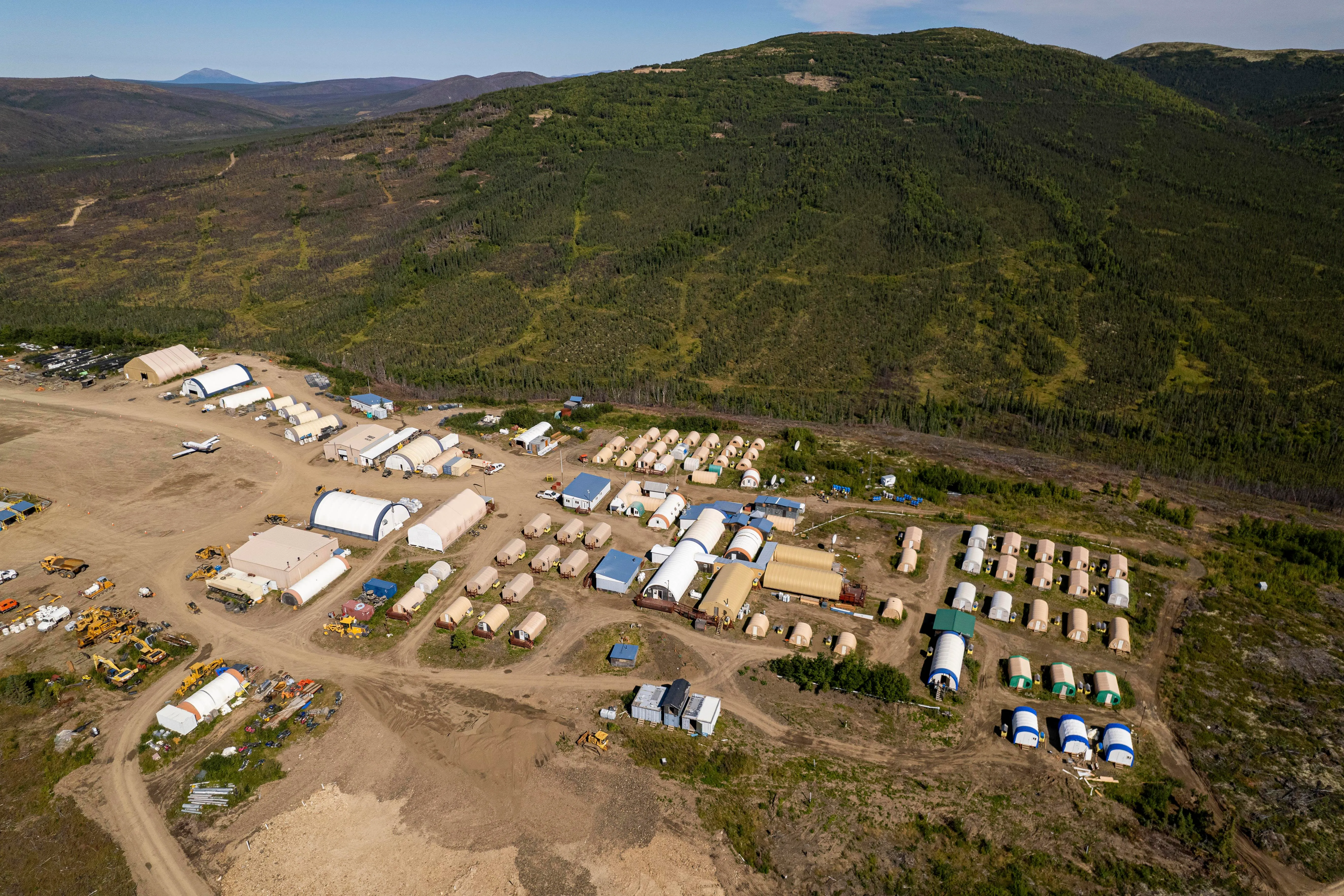 An overhead view of land cleared for many small buildings and storage sheds near a forested mountain.