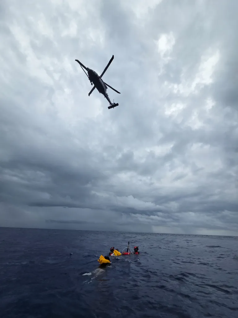 A helicopter hovers over three people in life rafts in the ocean on a cloudy day.