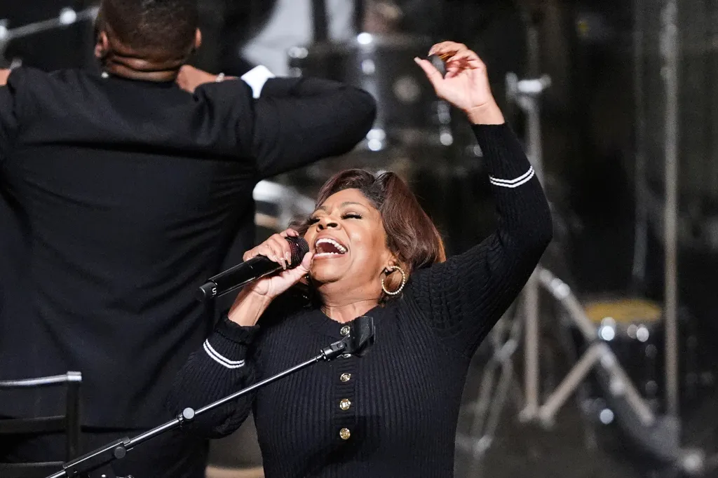 A woman in a black top sings into a microphone, with a man in a black suit partially visible behind her.