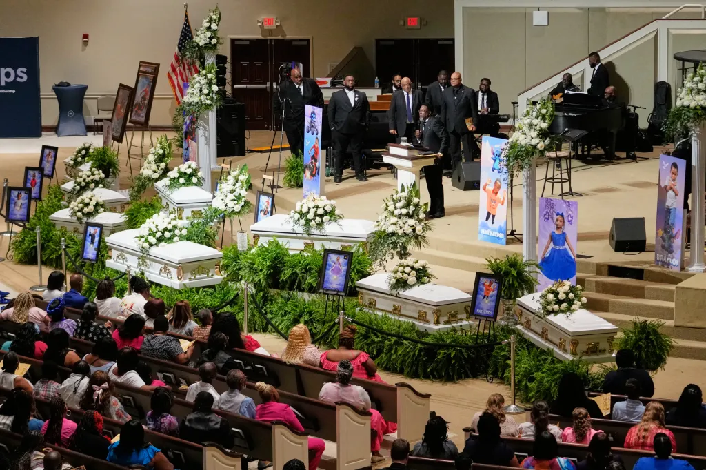 Six white caskets with white flowers on top are arranged at a funeral service, each accompanied by a child's portrait, with attendees seated in pews.