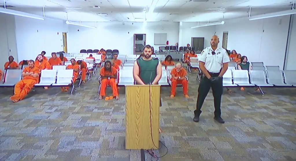 A man in an olive-green vest stands at a wooden podium, surrounded by other men in orange jumpsuits sitting on benches, and a sheriff's deputy standing to the right.