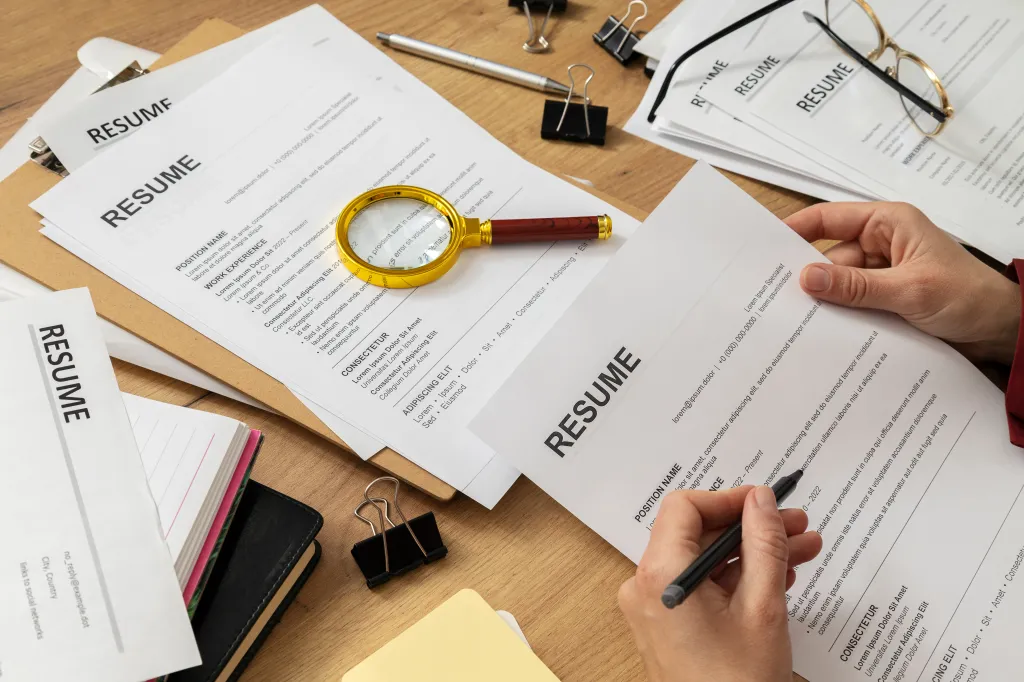 A person reviewing a resume, with other resumes, office supplies, and a magnifying glass on a wooden desk.