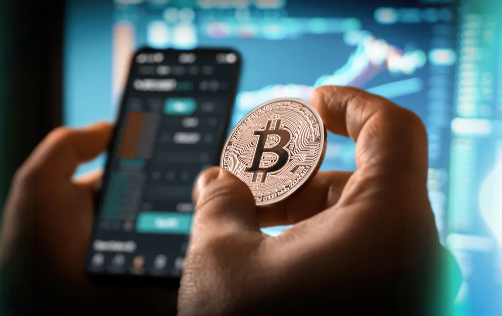 A person holds a Bitcoin coin while looking at a cryptocurrency trading app on a smartphone, with charts and graphs on a blurred screen in the background.