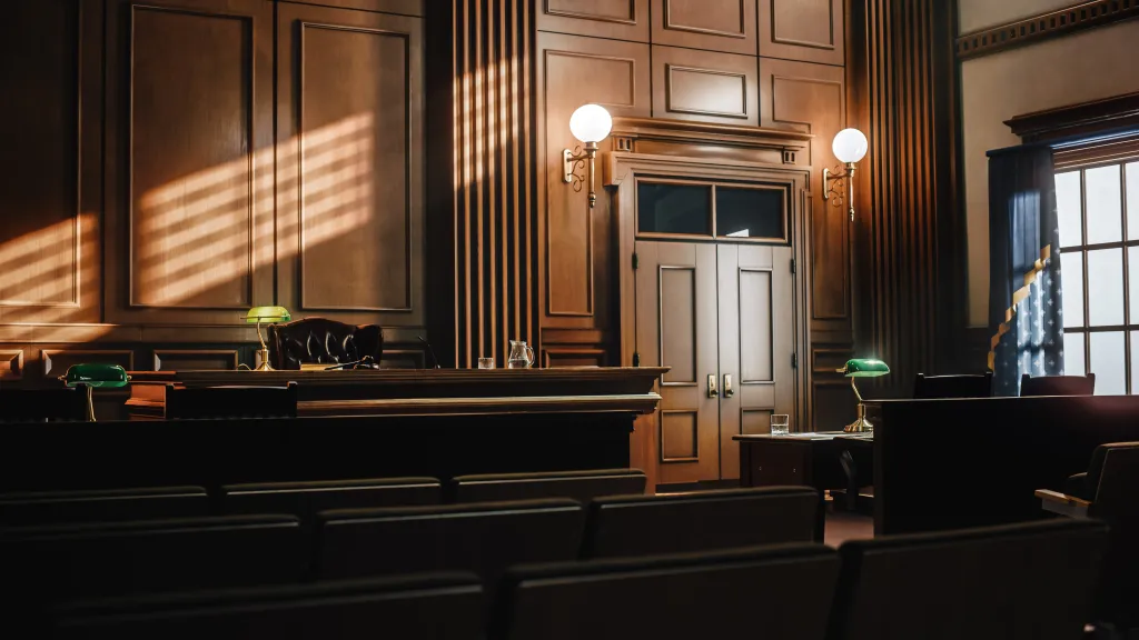 Illustration of an empty American-style courtroom with wooden paneling and seating, featuring a judge's bench, tables for the defense and plaintiff, and large windows.