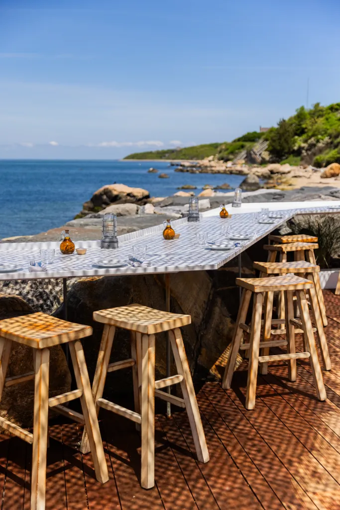 Outdoor dining area with a long table and wooden stools overlooking the ocean.