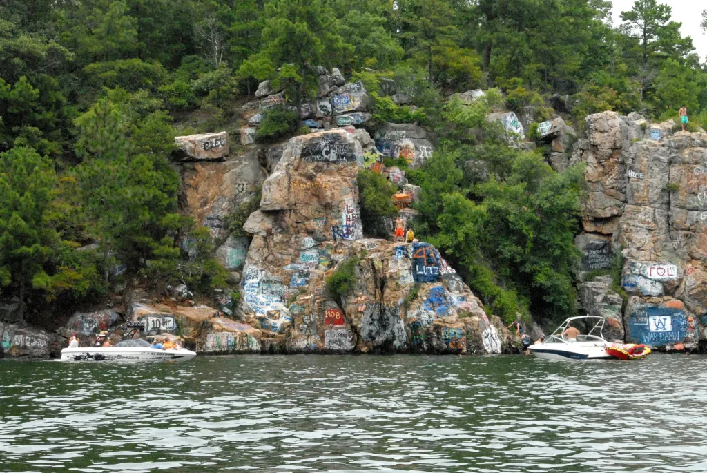 People gathered on boats and a rocky cliff face covered in graffiti at Chimney Rock, AL.