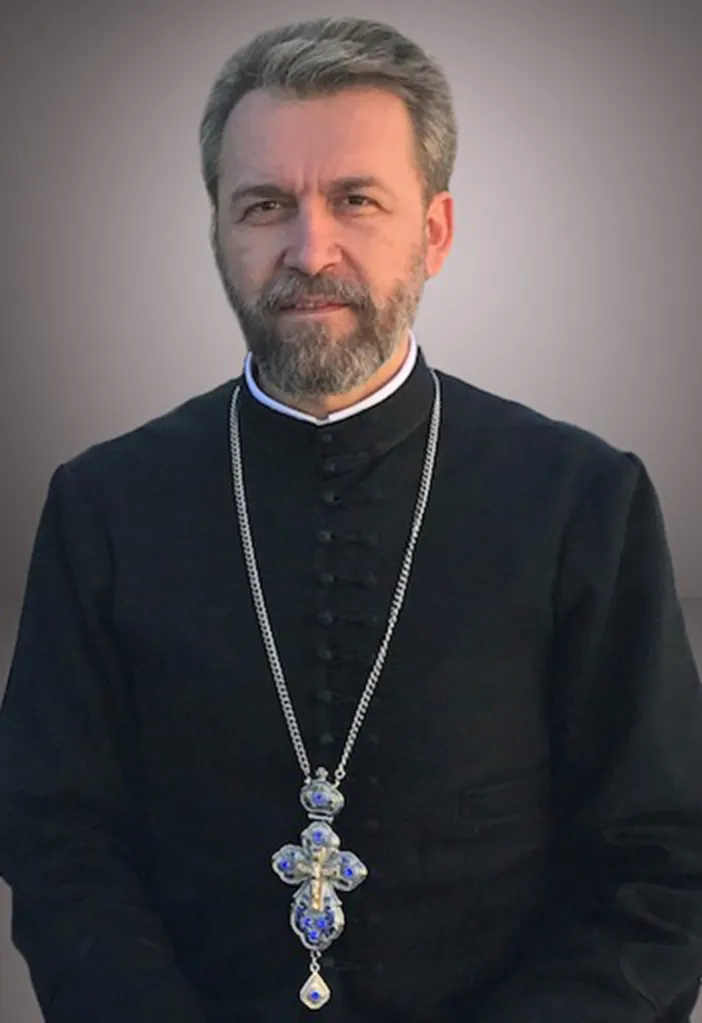 A bearded priest in a black cassock and white collar, wearing a silver chain with a blue ornate cross.