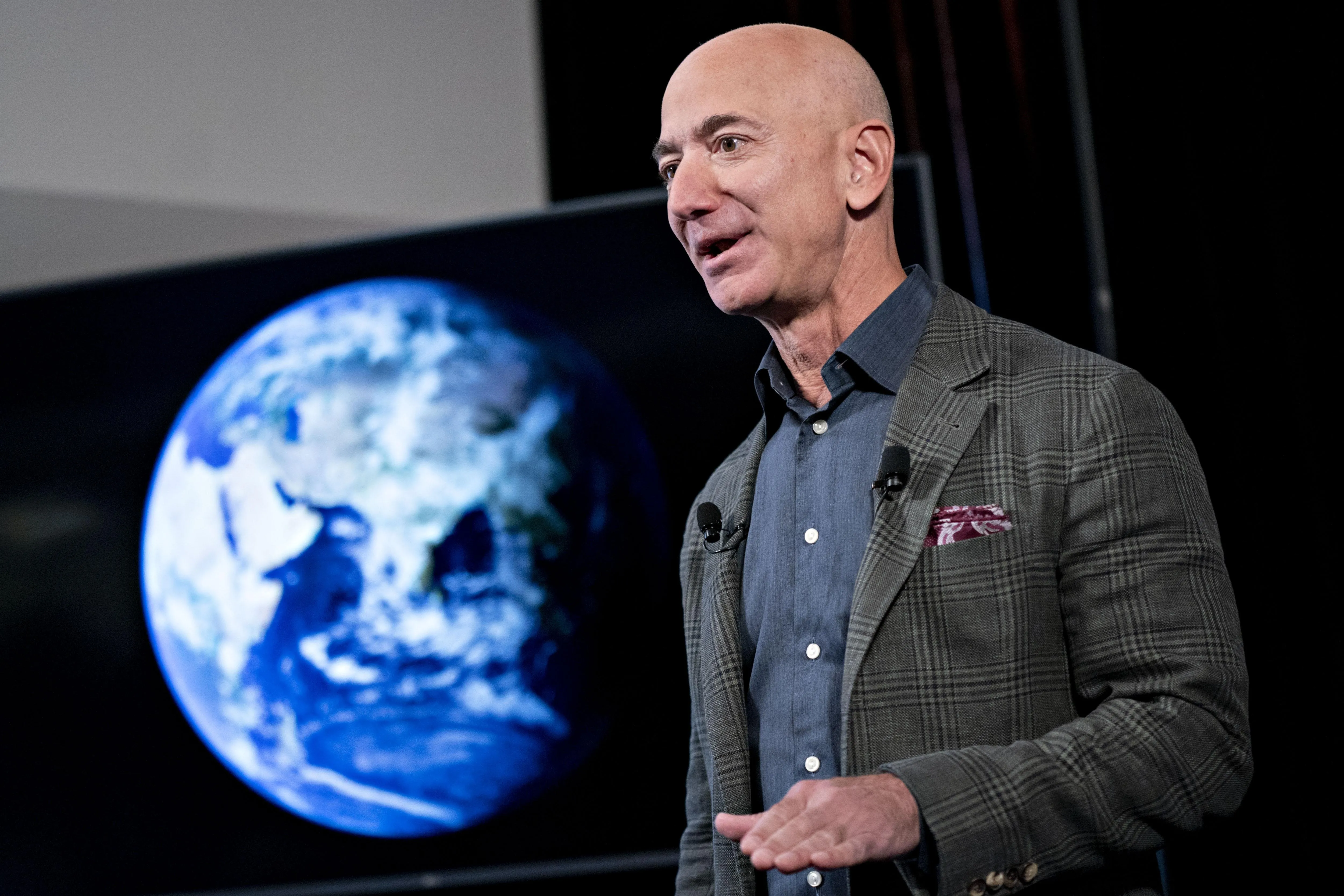 Jeff Bezos, in a gray jacket and blue shirt, standing near a large screen showing the planet Earth.