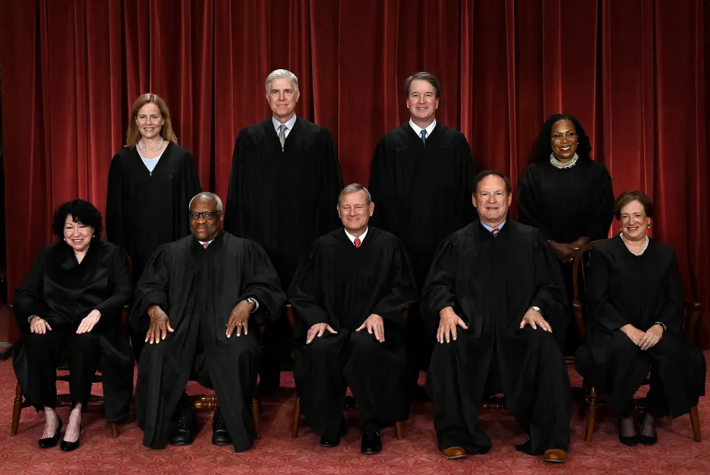 Nine Supreme Court justices, four women and five men, pose in black robes against a red curtain.