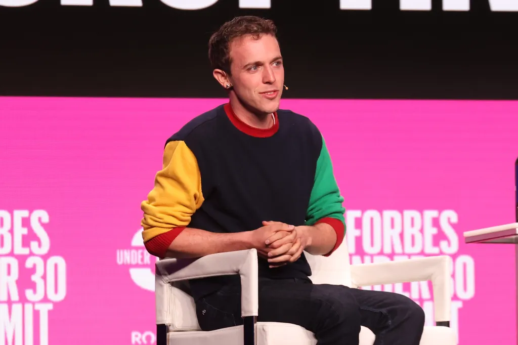 A young man with light brown hair and a colorful sweatshirt speaks from a white chair at the Forbes Under 30 Summit.