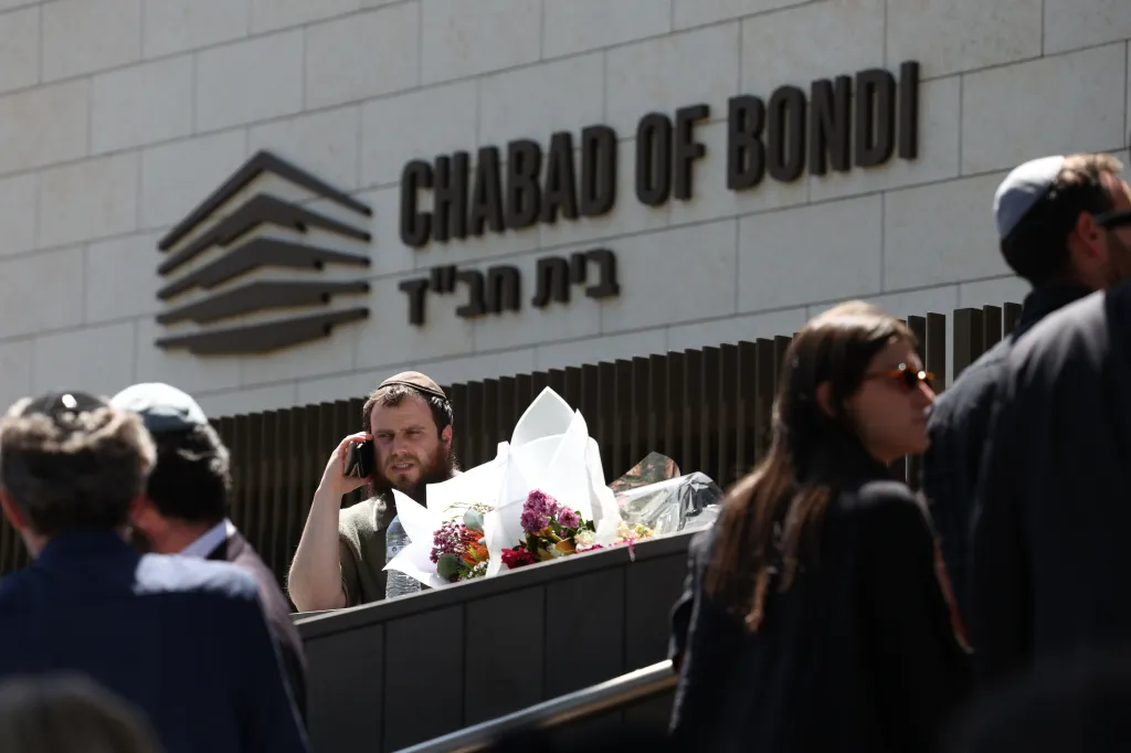 A man with a beard and a kippah talks on a phone while holding flowers in front of the Chabad of Bondi building.