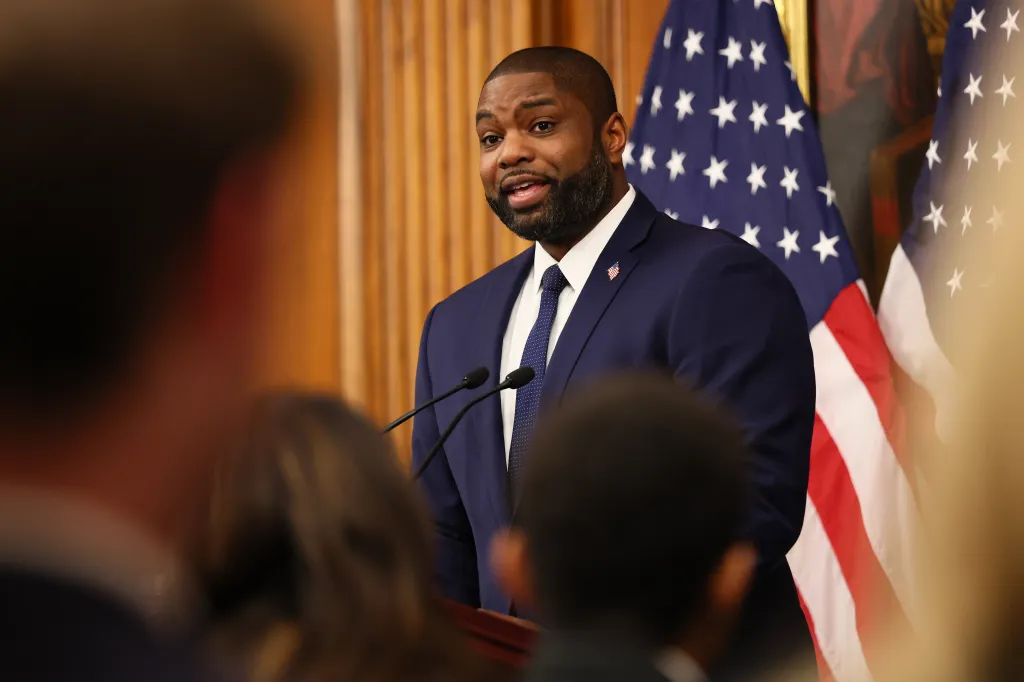 Byron Donalds speaking at a podium with two American flags behind him.