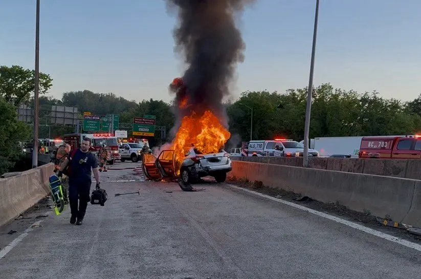A car engulfed in flames with thick black smoke rising from it on a highway, with emergency vehicles and personnel around.