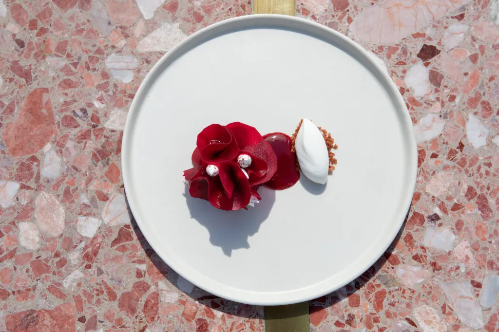 A dish from Jeune et Jolie restaurant, featuring red petals, white spheres, red sauce, and a quenelle of white sorbet on a white plate.