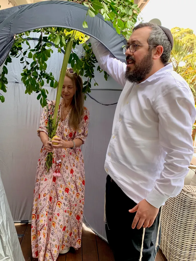 Nikki Eli holding an etrog and lulav inside a sukkah with Rabbi Eli Schlanger, possibly during Sukkot.