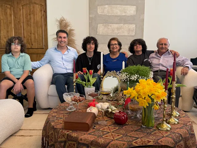A family of six, including children, parents, and grandparents, sitting together with a Haft-Seen table for Persian New Year (Nowruz) in the foreground.
