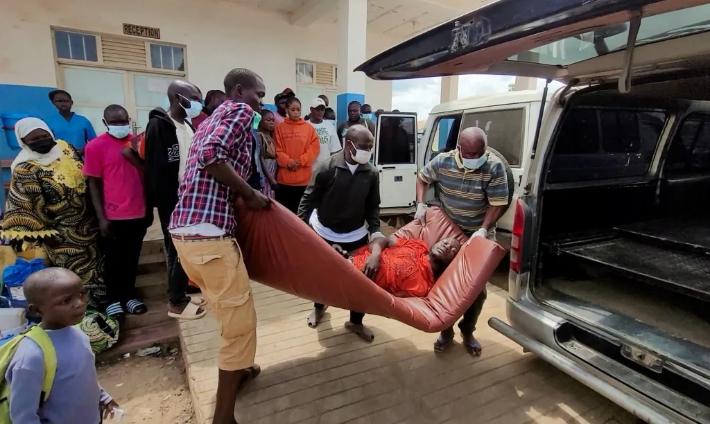 People carry a patient on a stretcher toward an ambulance as others look on outside a building with a 