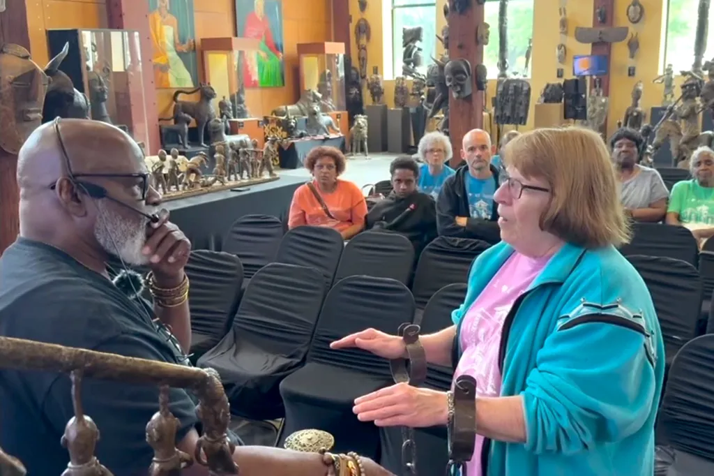 Two people in the foreground discussing shackles, with an audience and sculptures in the background.