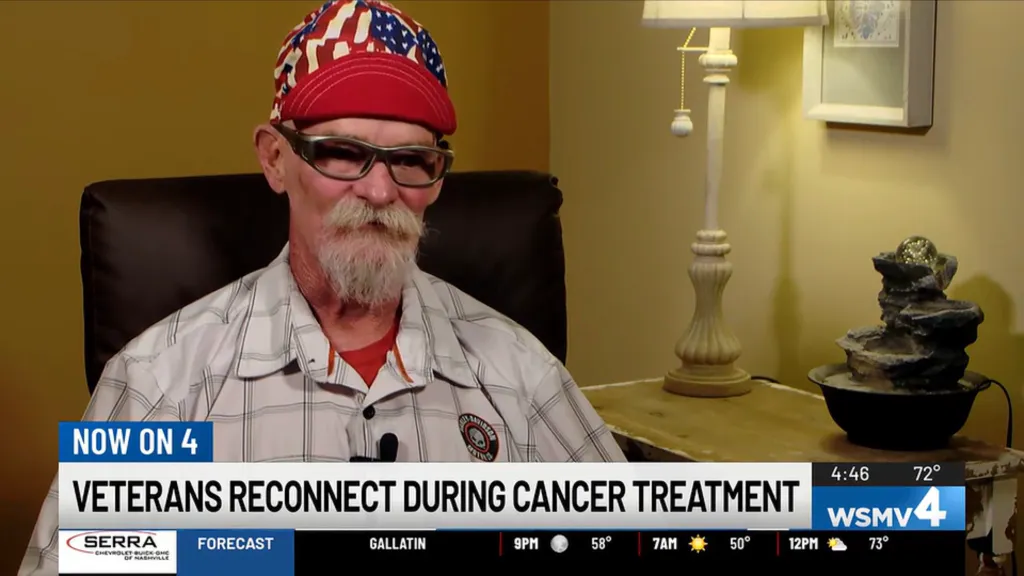 A veteran with a white beard, wearing an American flag-patterned hat and sunglasses, is interviewed on a news channel about veterans reconnecting during cancer treatment.