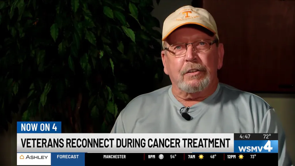 A man with glasses and a baseball cap is shown on a TV screen with the headline 
