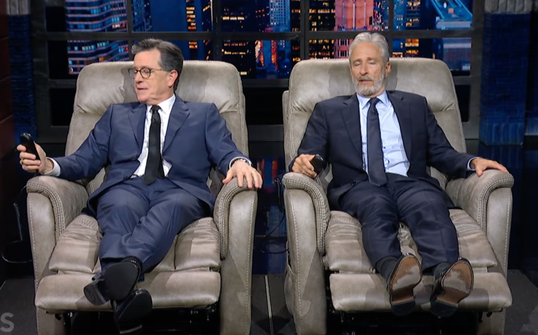 Stephen Colbert and Jon Stewart relaxing in recliners on a TV set.