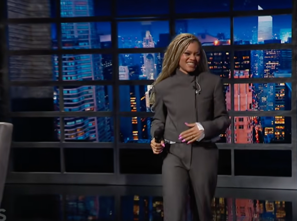 A smiling woman with braided hair wearing a dark gray suit and holding a microphone stands before a window overlooking a cityscape at night.