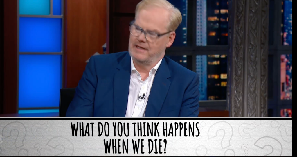 Comedian Jim Gaffigan in a blue suit and white shirt, talking on a talk show set, with the question