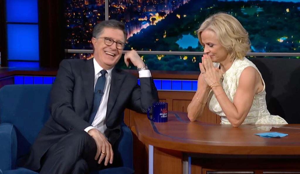 Stephen Colbert laughing with his hand on his chin, sitting next to a woman with hands clasped as if in prayer.
