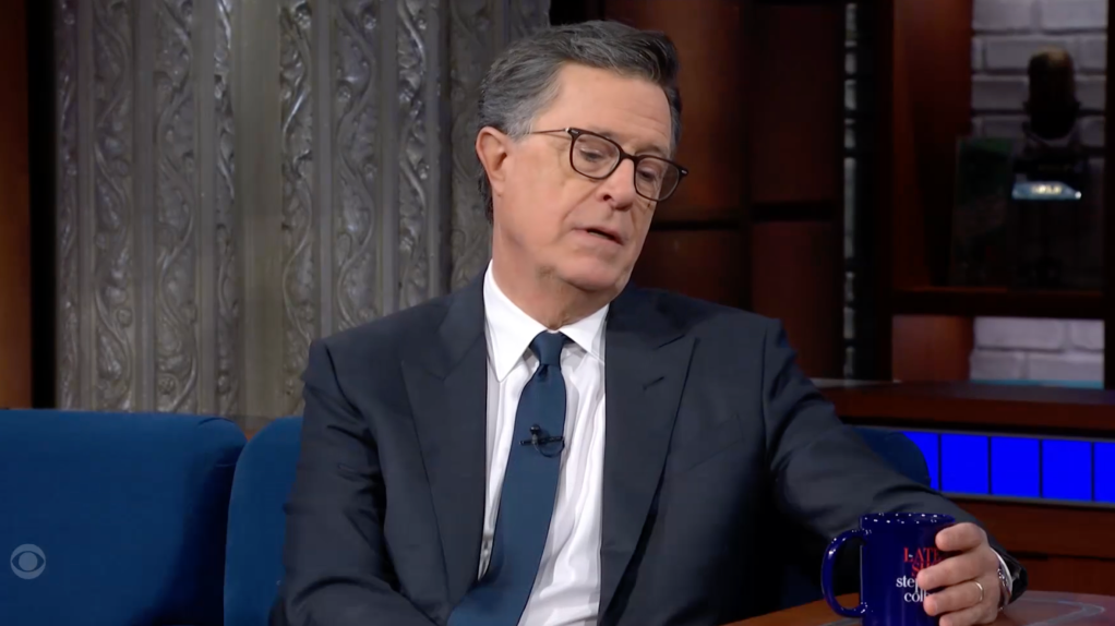 Stephen Colbert wearing a suit and glasses, holding a blue mug, sitting on a blue couch in a television studio.