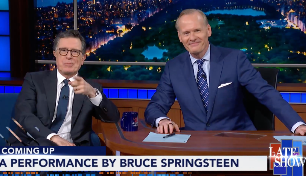 Stephen Colbert pointing at the viewer next to Jeff Glor at The Late Show desk.