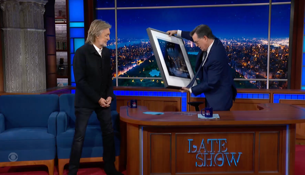 Paul McCartney smiling as Stephen Colbert holds up a framed photo on the Late Show set.