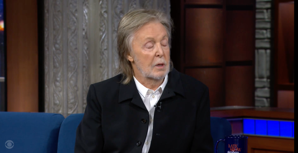 Paul McCartney sitting on a blue couch, wearing a black jacket over a white shirt, with a Late Show with Stephen Colbert mug in front of him.