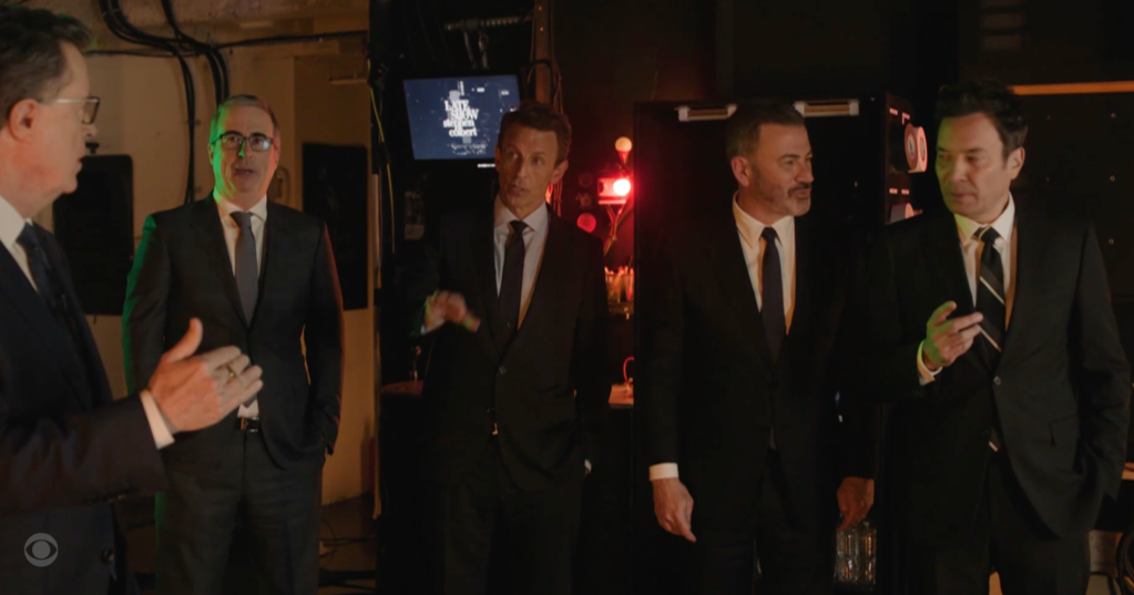 Five late-night hosts, from left to right, Colbert, Oliver, Meyers, Kimmel, and Fallon, standing in suits.