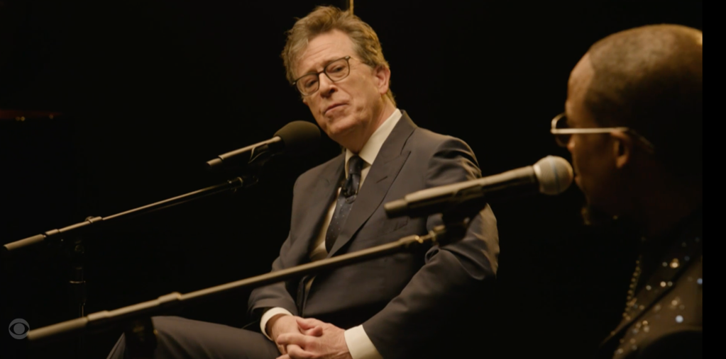 Stephen Colbert in a suit and glasses, speaking into a microphone.