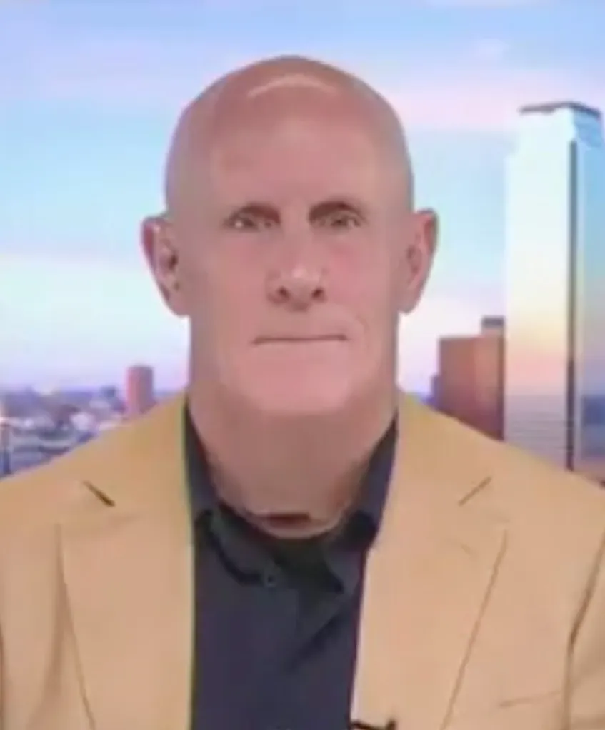 A bald man in a tan jacket and dark shirt, with a city skyline in the background.