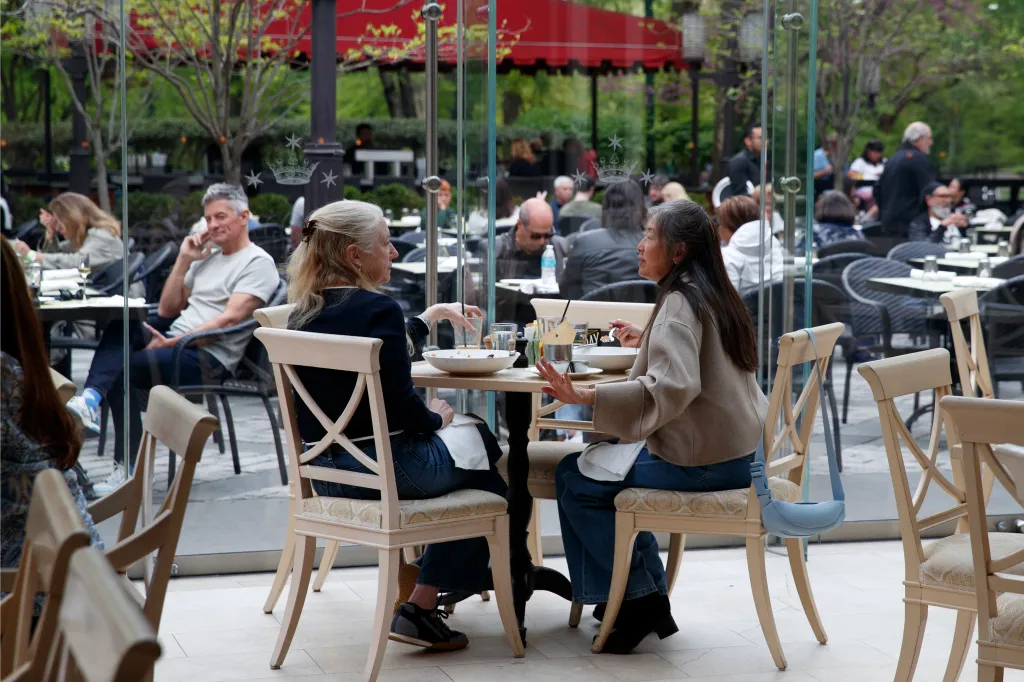Two women sit at a table inside a restaurant with large glass windows, conversing.