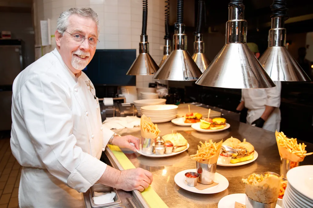 A smiling chef in a white uniform standing in a kitchen with plates of burgers and fries on a counter.